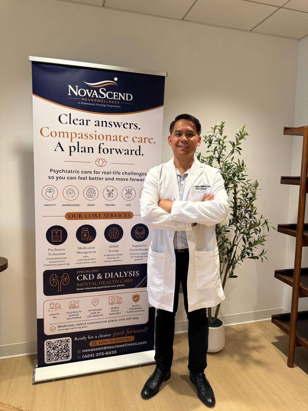 A man in a white coat standing with arms crossed next to a tall informational banner for Nova Sound Neurowellness, which offers psychiatric care services. The man is smiling and standing indoors near a plant and a wooden shelf.