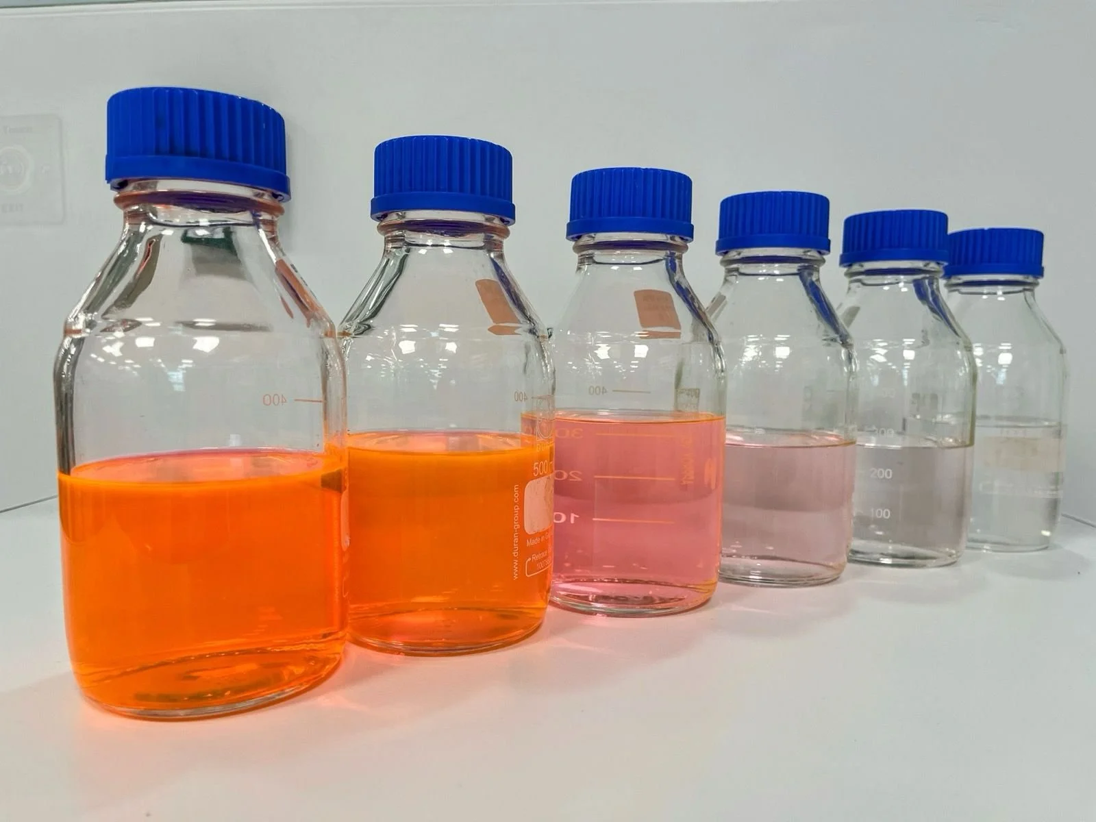 Six clear glass bottles with blue caps containing varying levels of orange and pink liquids, arranged in a row on a white surface.