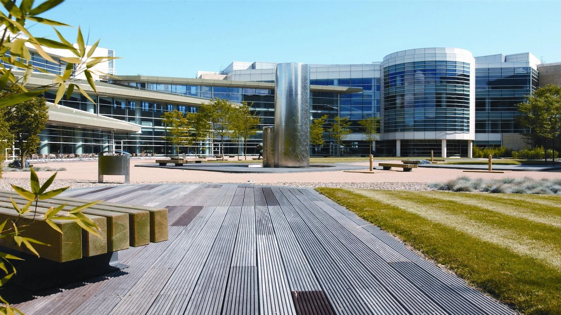 Modern office building with curved glass facade, surrounded by trees, benches, and a water fountain in the foreground. Building 500, Discovery Park, DP, Sandwich, Kent, Business park, Science park