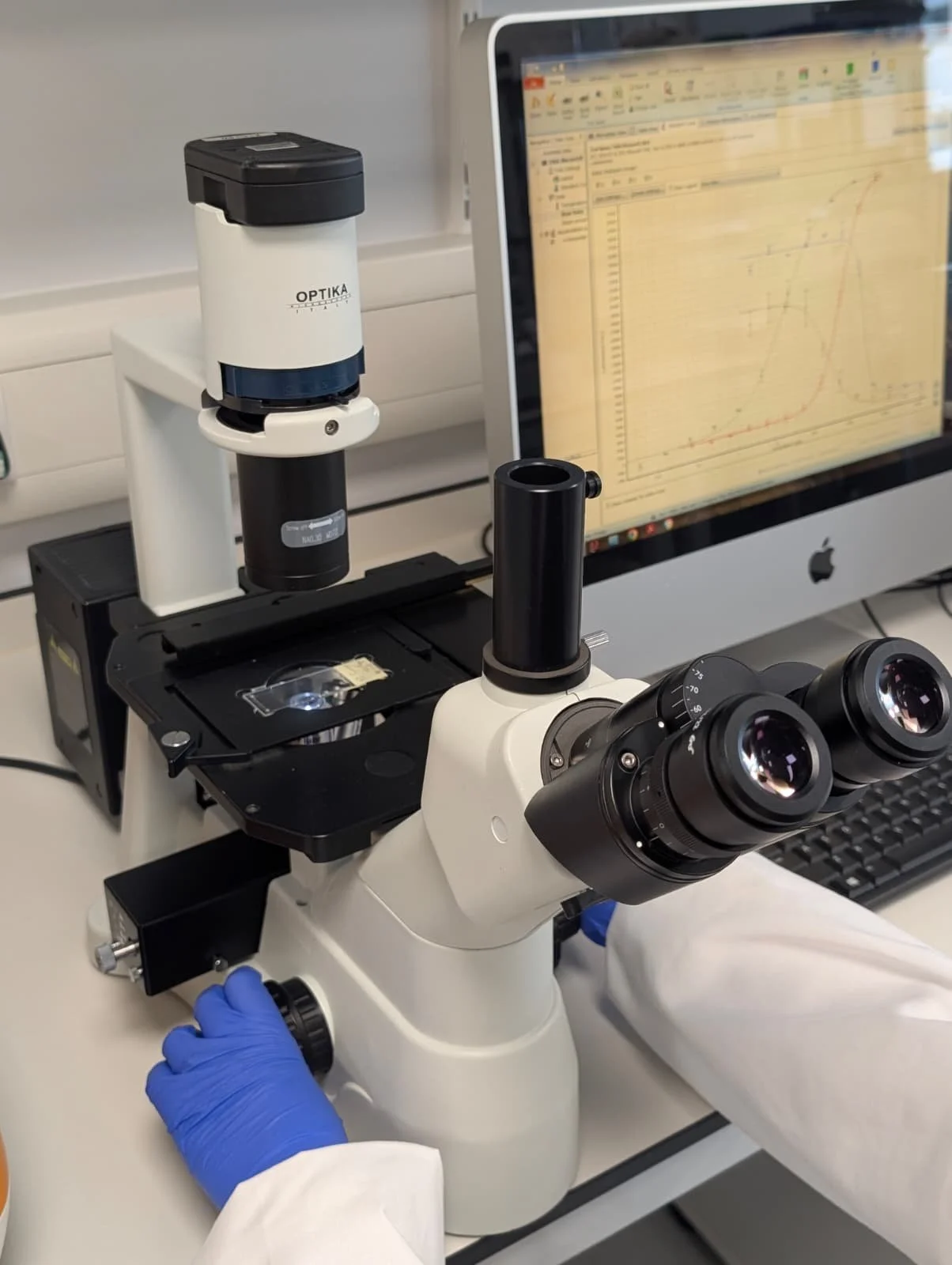 A laboratory scientist using a microscope with lenses and a computer screen displaying a graph in the background.