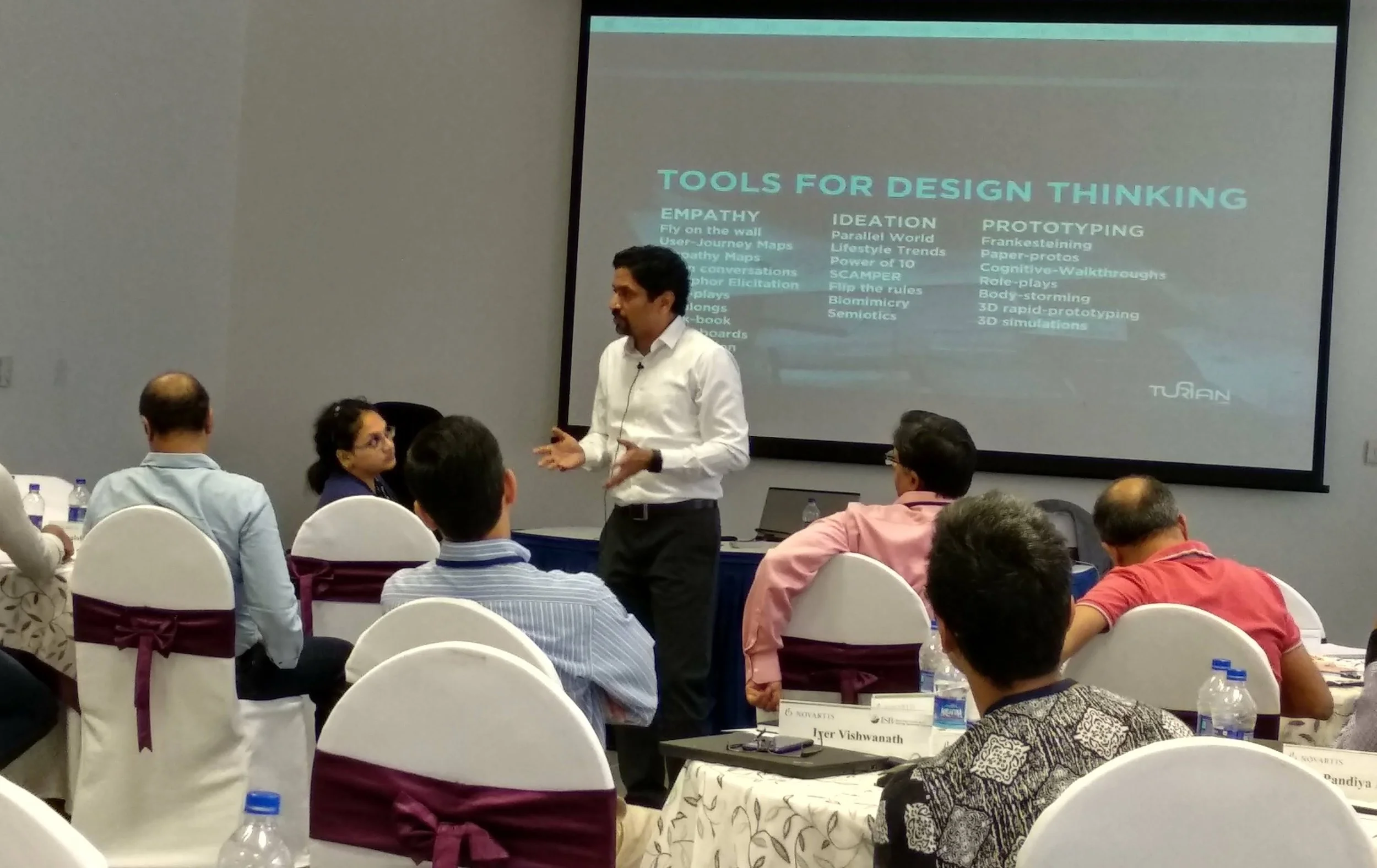 A man in a white shirt giving a presentation at a conference. Several people seated at tables listen, with a large screen behind him displaying a slide titled 'Tools for Design Thinking' with categories such as Empathy, Ideation, and Prototyping.