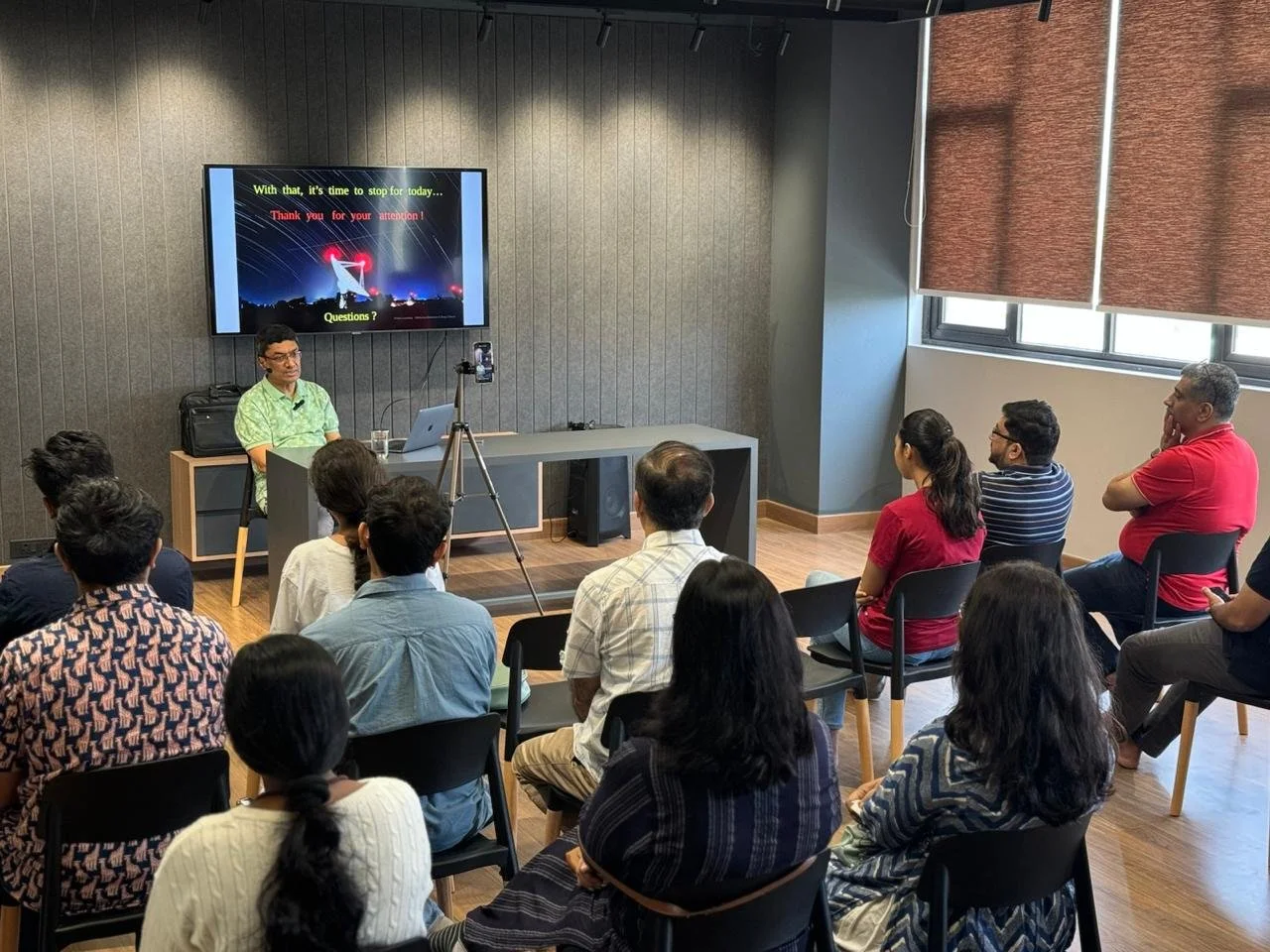 A man giving a presentation to a group of people in a conference room with a large screen displaying a slide and a camera on a tripod capturing the session.