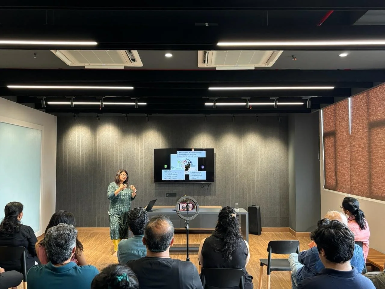 A woman giving a presentation in a conference room with a group of people seated and a screen behind her displaying slides.