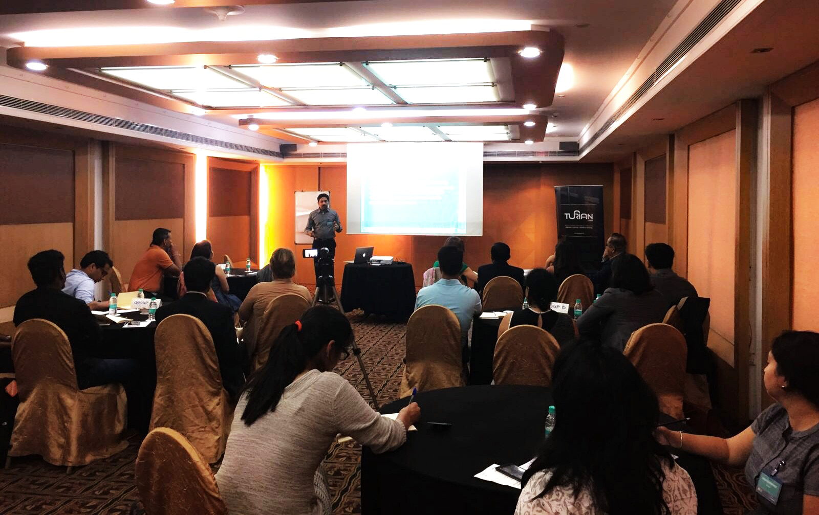 A conference room with attendees sitting at round tables listening to a speaker at a presentation. Megatrends. Foresight. Strategy. 2019