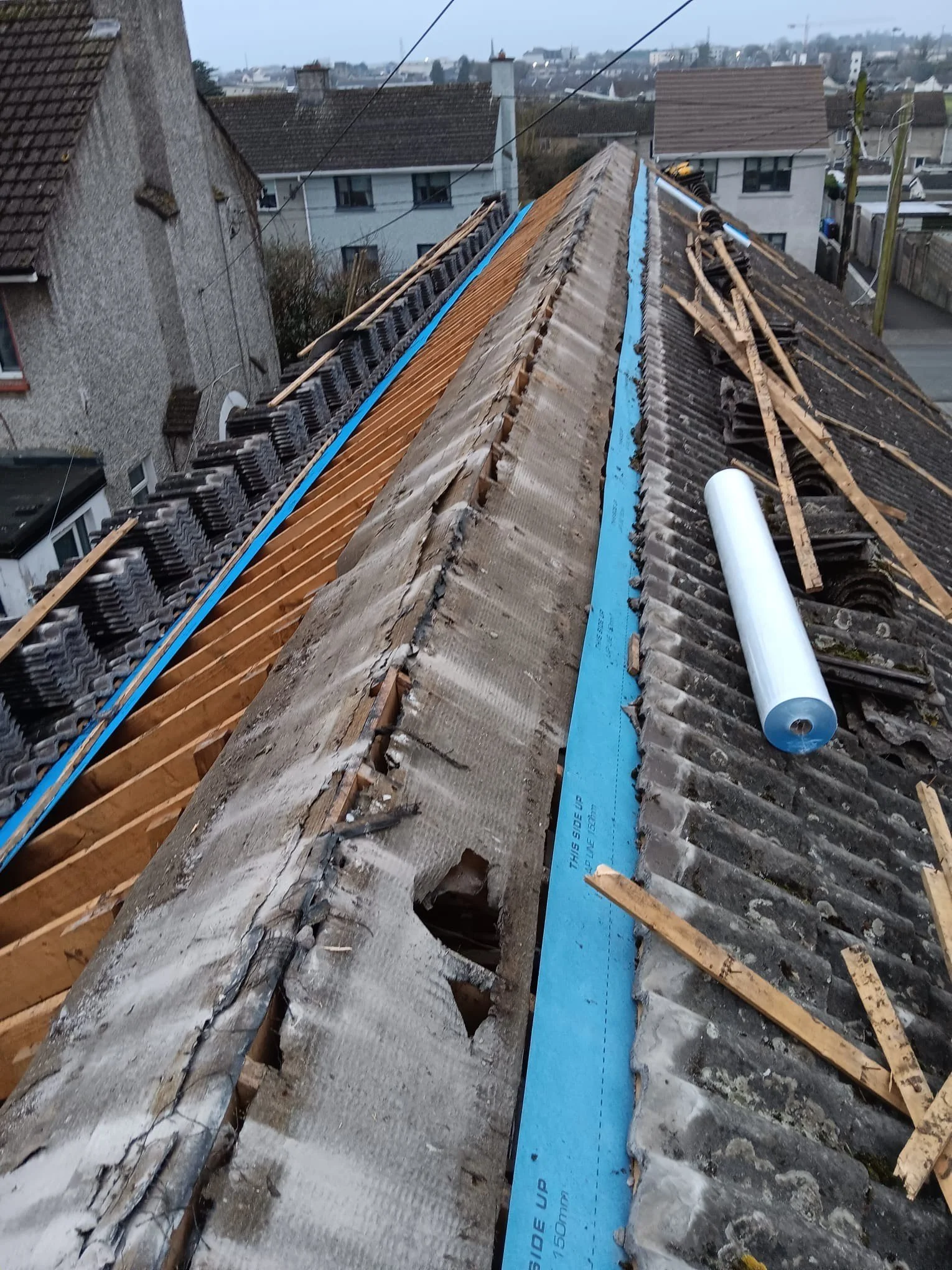 Roof of a building undergoing repair with old and new tiles, wooden beams, blue insulation strip, and construction tools, in a residential neighborhood.