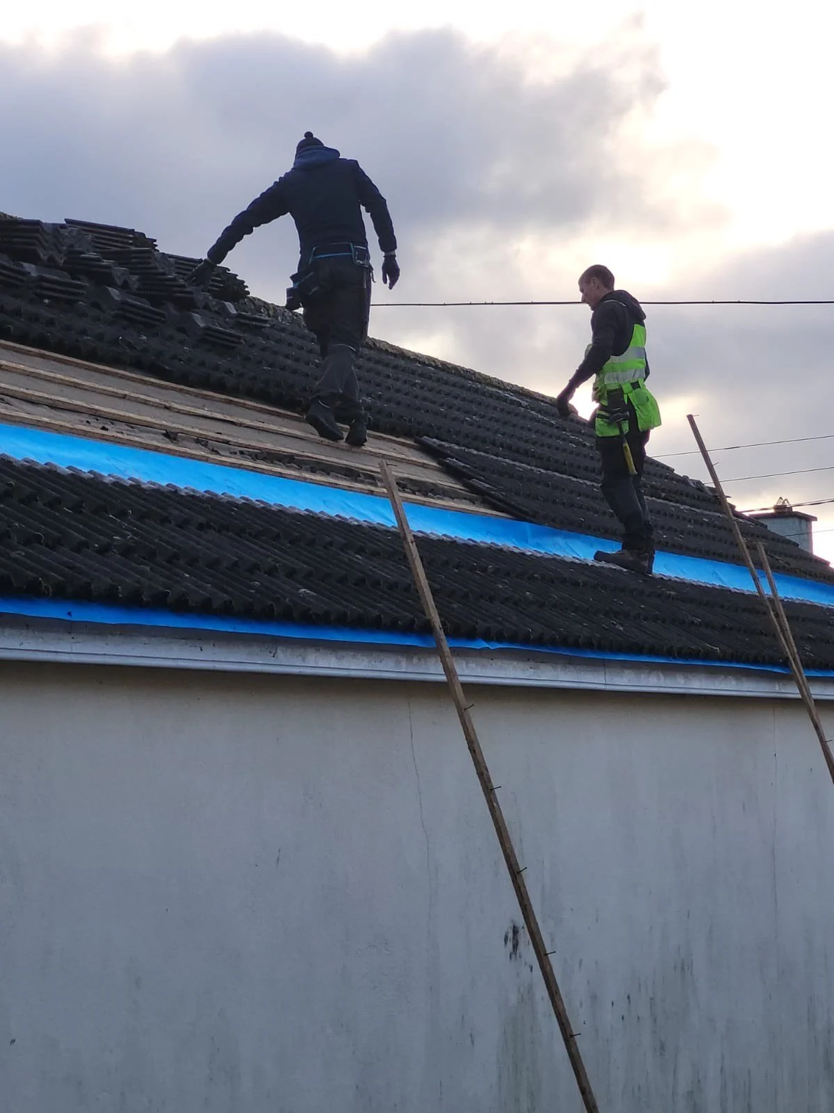 Two workers on a sloped roof performing repairs or maintenance, with part of the roof covered in black tiles and a section wrapped in blue material, set against a cloudy sky.