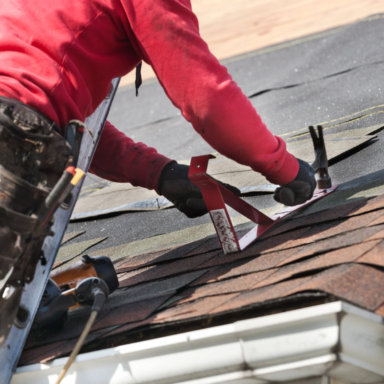A worker in red clothing installing asphalt shingles on a roof, using a ruler and hammer.