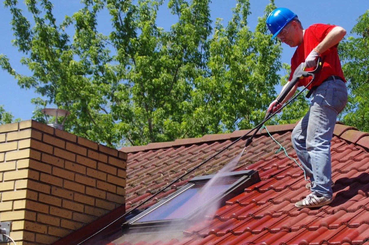 A man wearing a red shirt, blue jeans, white sneakers, and a blue safety helmet is cleaning a skylight on a red-tiled roof with a pressure washer.