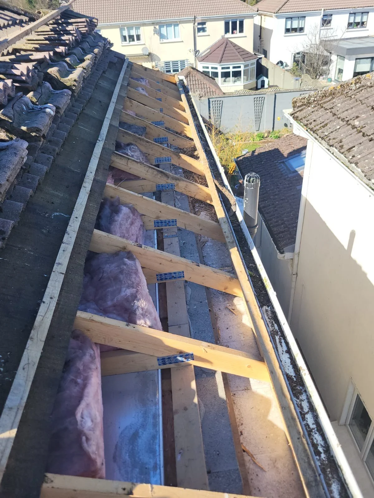 View of a house roof with old tiles removed, showing wooden roof framework and insulation material, with neighboring houses in the background.