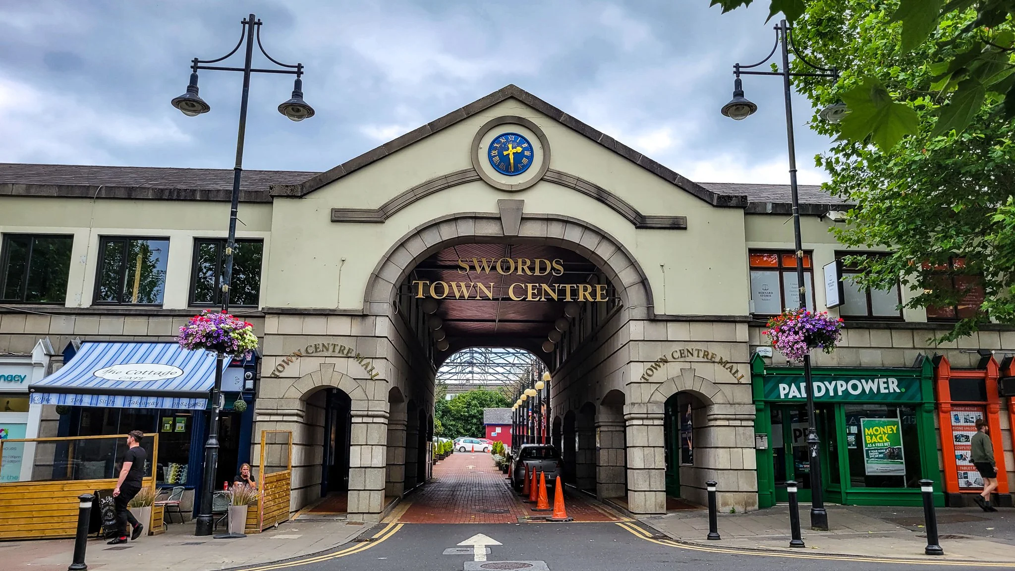 The entrance to Swords Town Centre with an archway, clock with a cross on it, and shops including a pad power store and a café with outdoor seating, trees, and street lights.