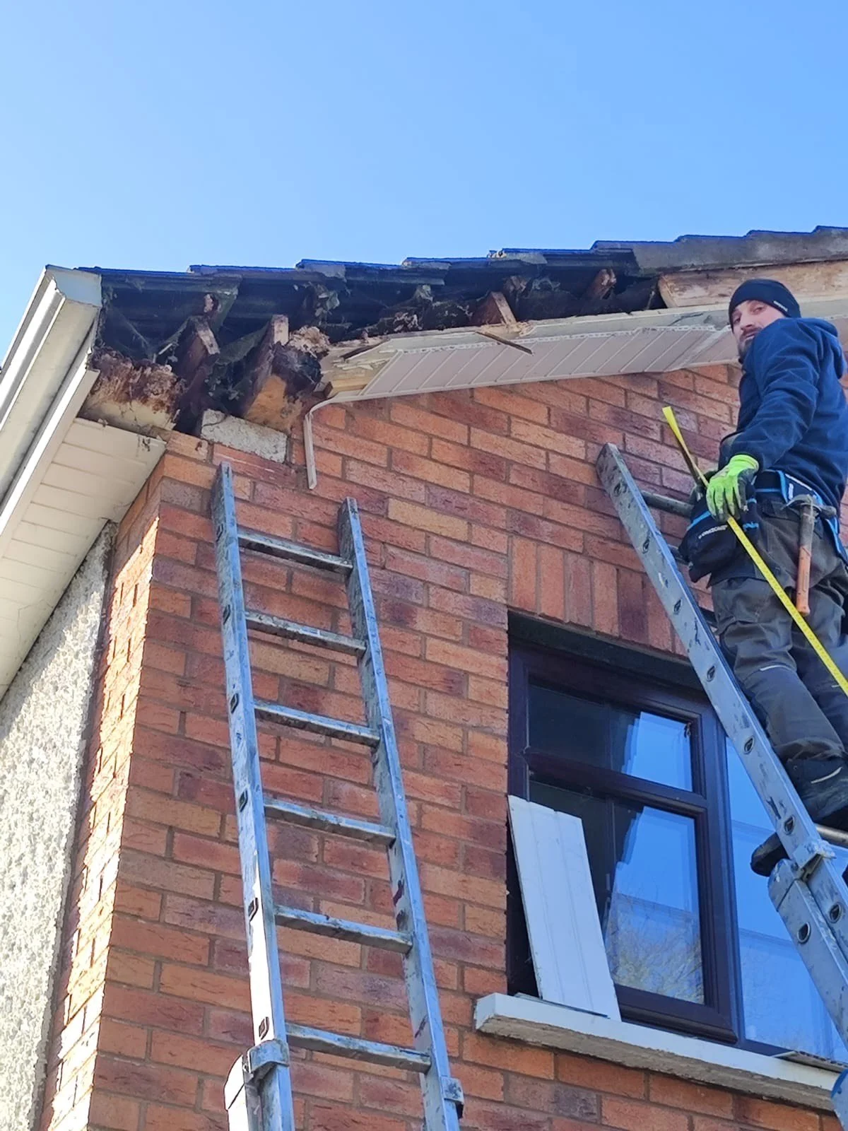 A worker is standing on a ladder fixing the gutters and soffit at the top corner of a brick house. The worker is wearing gloves, a beanie, and work clothes, holding a measuring tape, with another ladder leaning against the house.