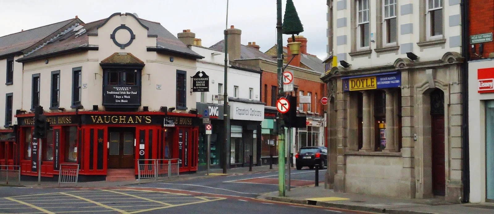 Street view showing Vaughans pub, a brick building labeled Doyle estate agents, a car, and various shopfronts with signs in an urban area.