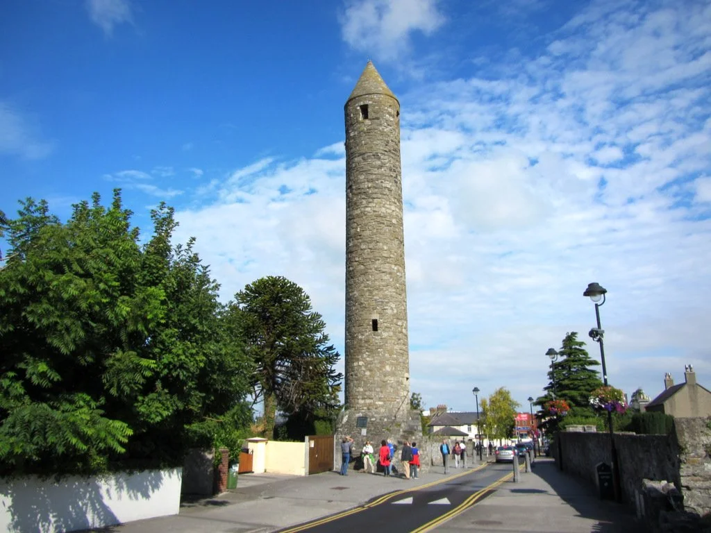 Stone round tower with conical roof in a small town, with people walking nearby and green trees under a blue sky with clouds.