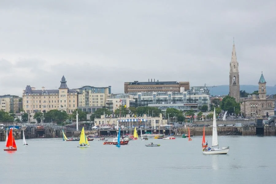 Sailboats with colorful sails on a body of water in front of a cityscape with buildings, church steeples, and mountains in the background under a cloudy sky.