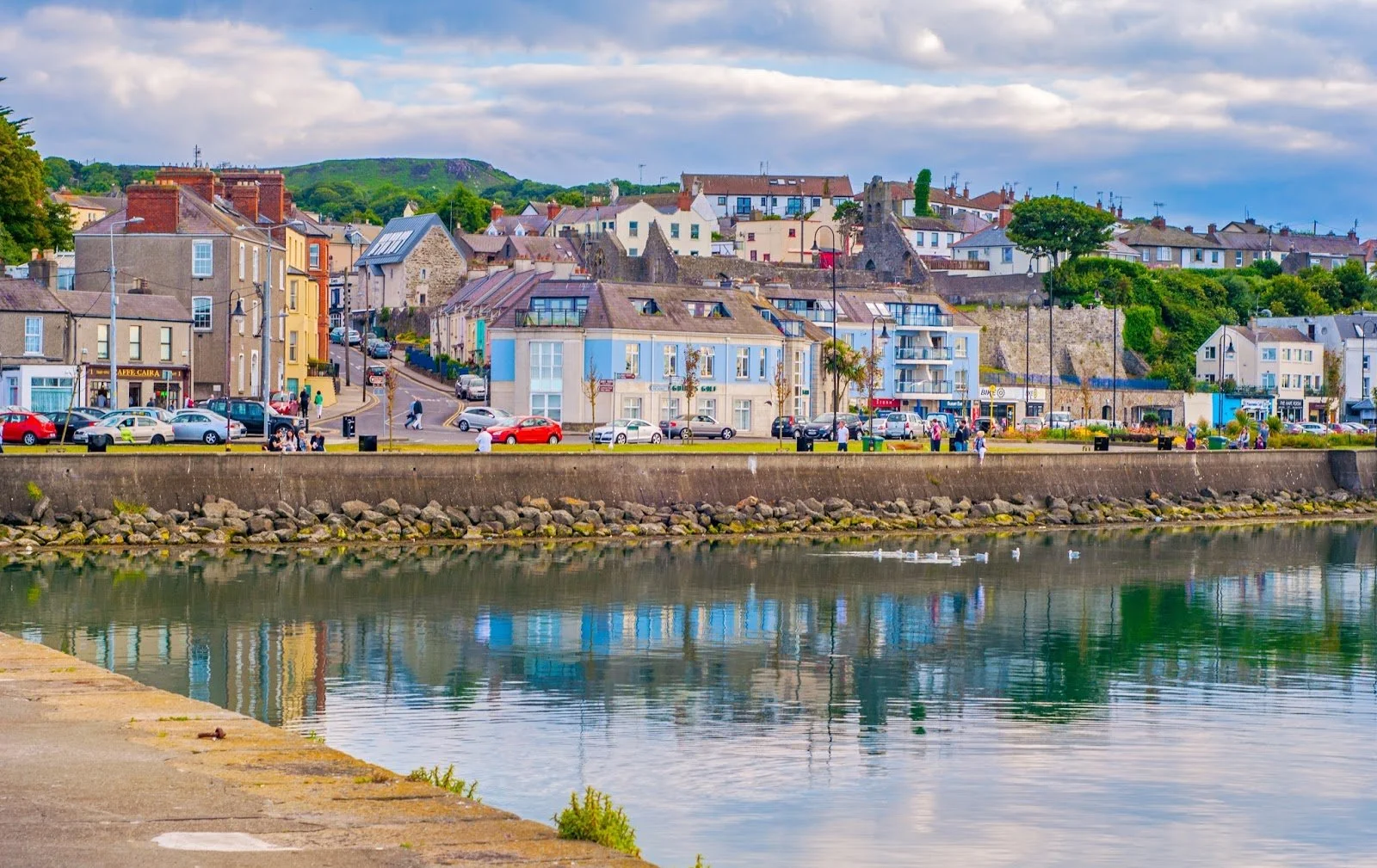 A colorful town with buildings along a waterfront, trees, parked cars, people walking, and a cloudy sky with hills in the background.