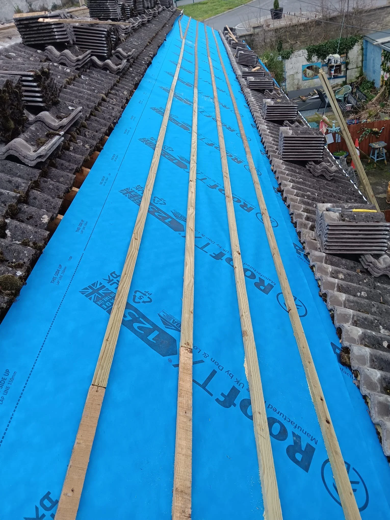 Roof under construction with wooden battens and a blue underlayment sheet, old roofing tiles on the sides, and construction tools visible.