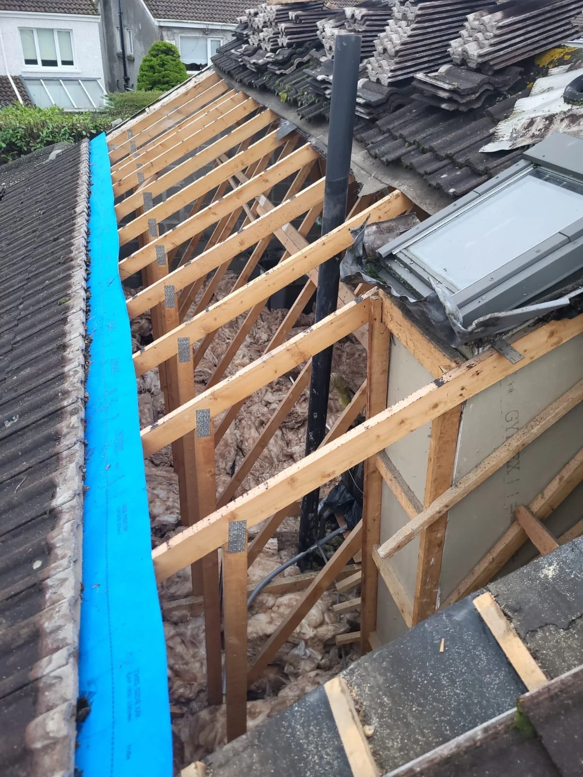 Roof under renovation showing wooden roof trusses, some roof tiles removed, and a skylight window.