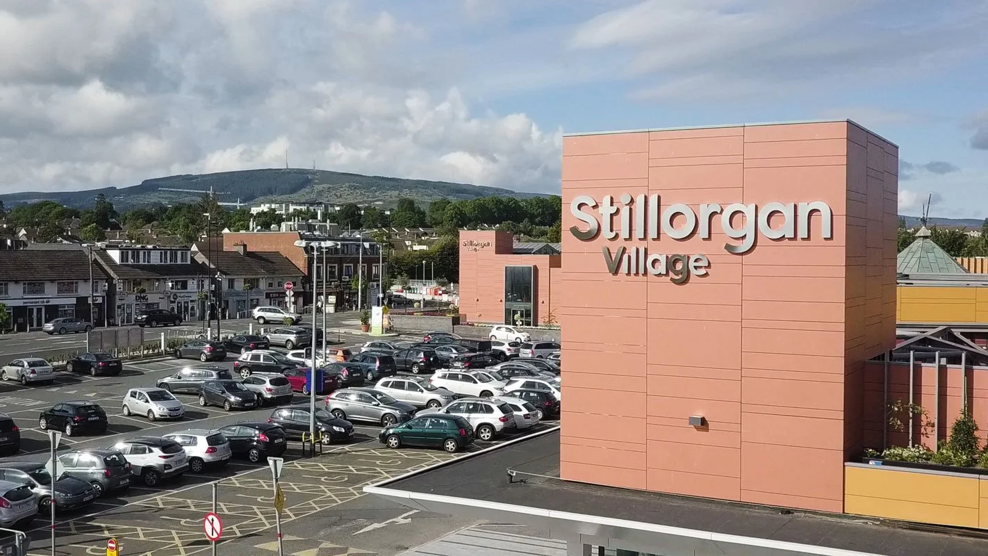 A parking lot filled with cars in front of a shopping center called Stillorgan Village, with a pink building bearing signage and a distant landscape with hills and partly cloudy skies.
