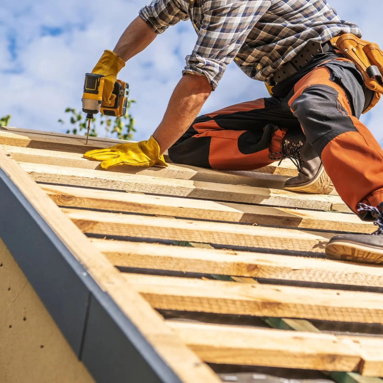 A person kneels on a wooden roof, using a power drill to work on the construction, wearing a plaid shirt, orange and black work pants, and yellow gloves.