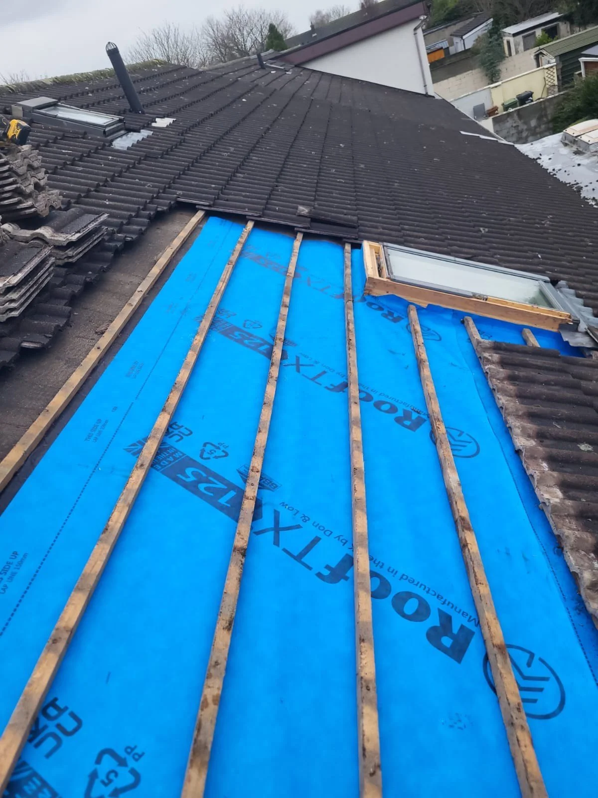 Roof under construction with blue roofing underlayment, wooden battens, and partially installed tiles, along with skylights visible.