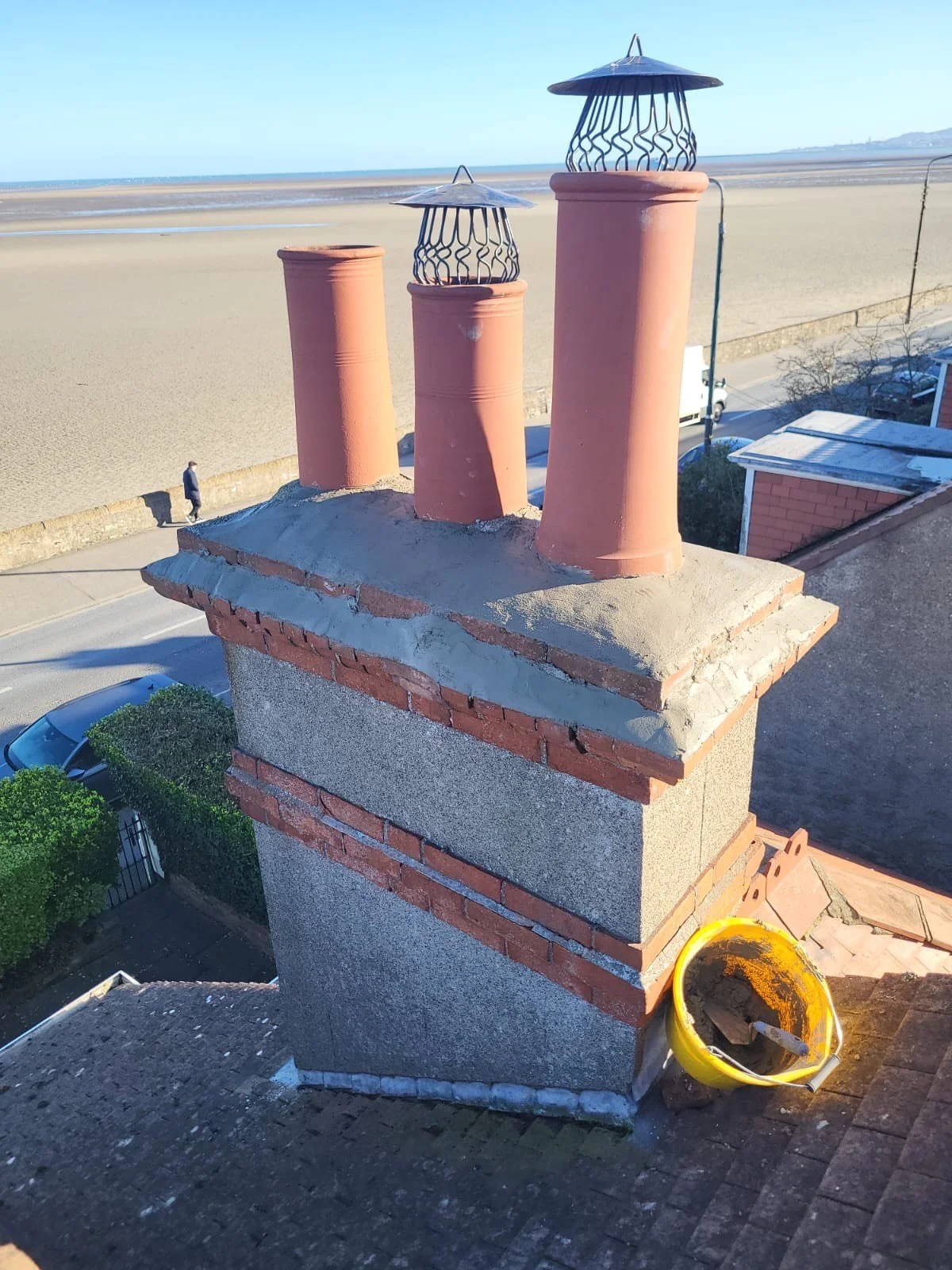 A rooftop chimney with three terracotta flues and metal caps under construction, with a yellow bucket and construction tools nearby, overlooking a sandy beach and ocean.