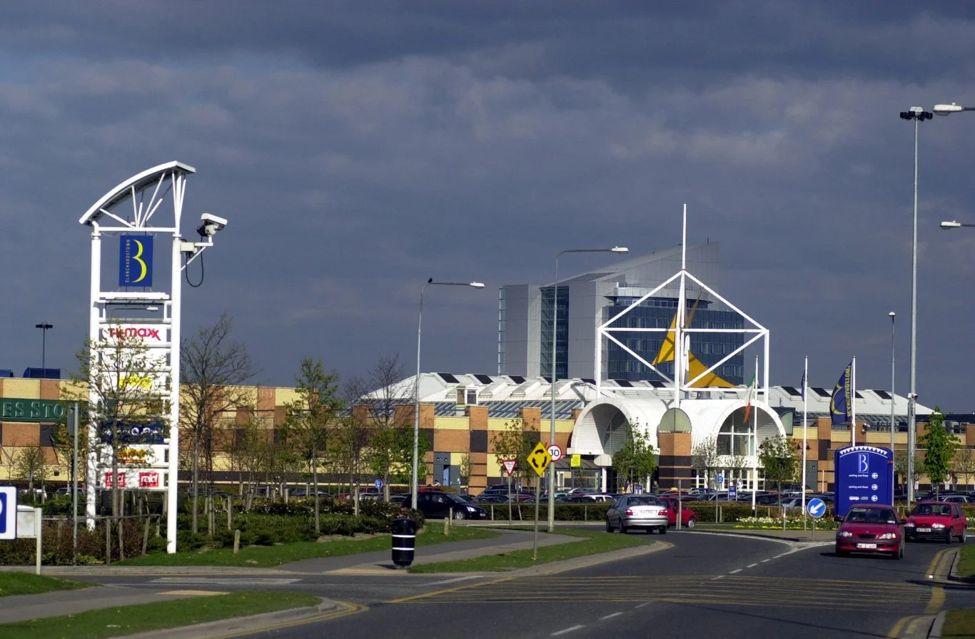 Shopping center with parking lot, cars, and traffic signs; large building with distinctive white framework and modern architecture, under a cloudy sky.