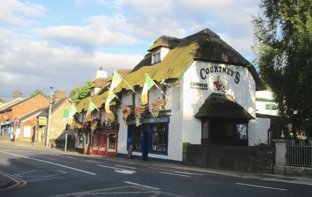A pub with a sign reading "Courtney's" on a white building with a thatched roof, surrounded by flowers and flags, on a street in a small town.