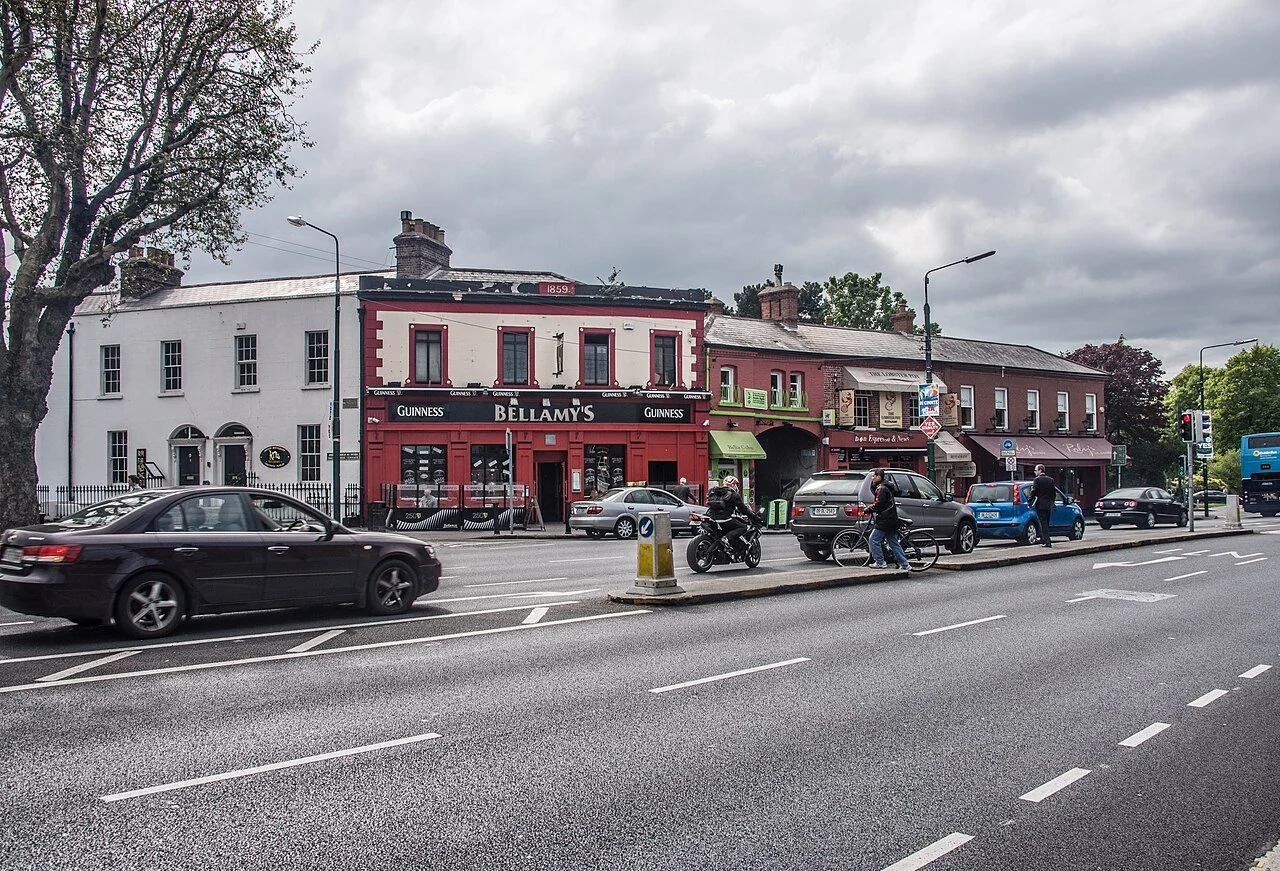 Street view showing cars and pedestrians in front of buildings with shops, including Bellamy's pub, on a cloudy day.
