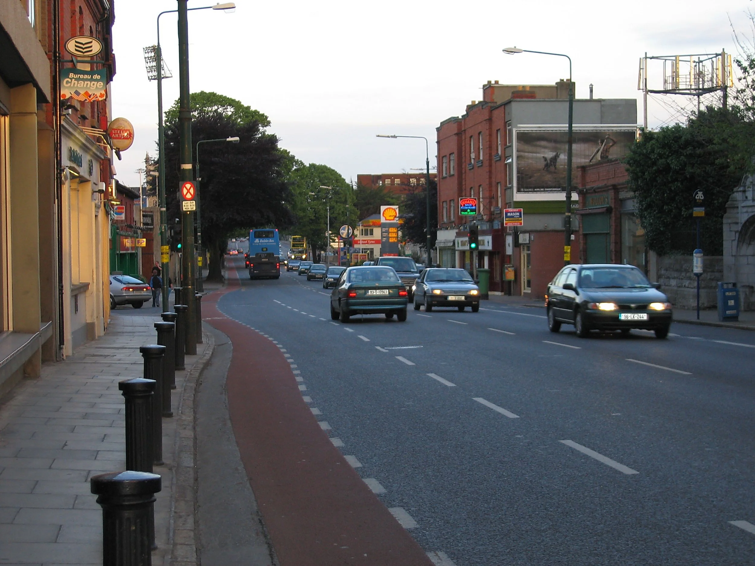 A busy city street with cars, a bus, and buildings on either side, with a Shell gas station sign visible in the background.