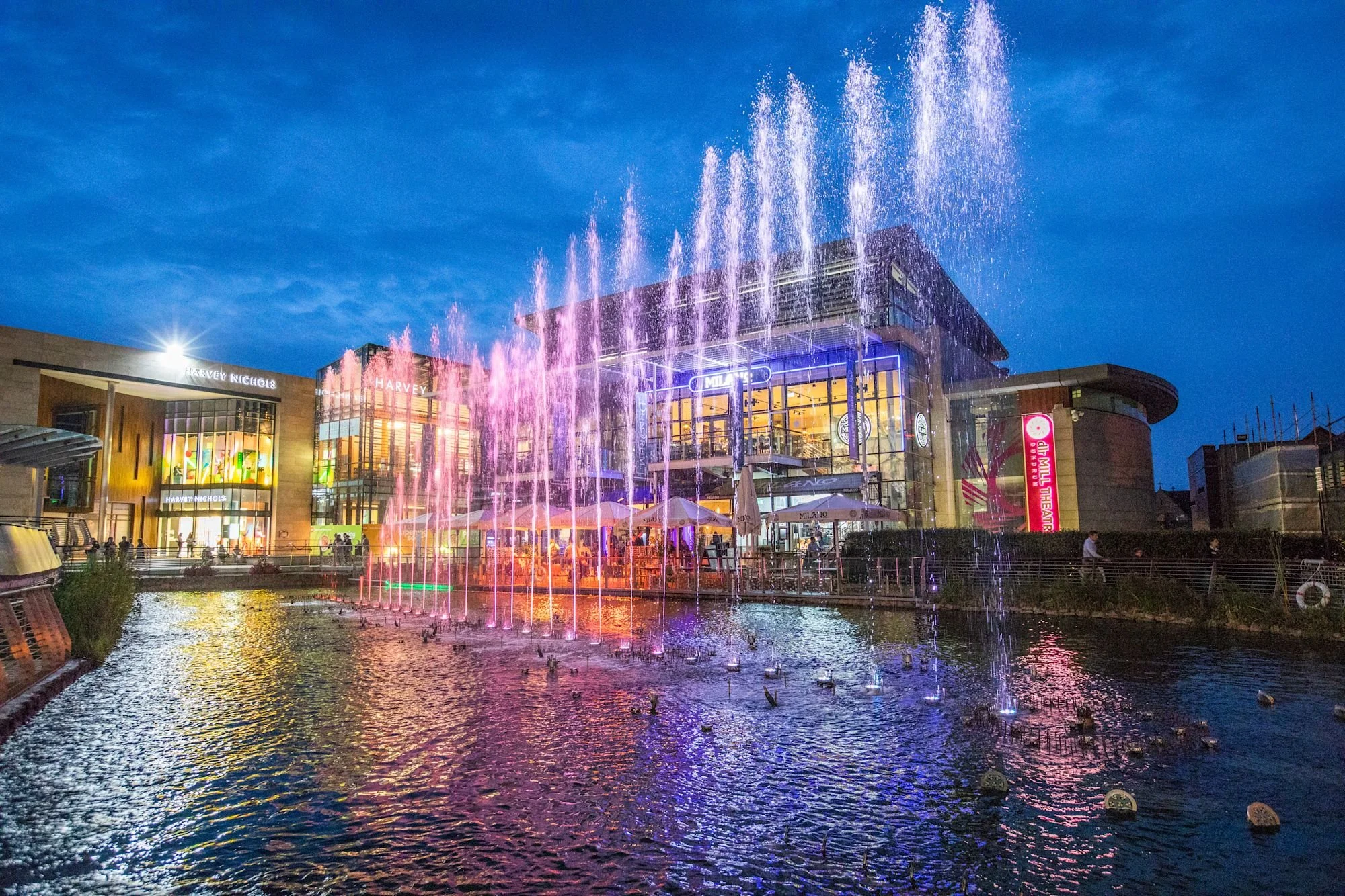 Colorful illuminated fountain with water jets shooting up, located in front of a modern shopping mall at dusk. The mall has large glass windows and neon signs, with people walking nearby and a canal reflecting the lights.
