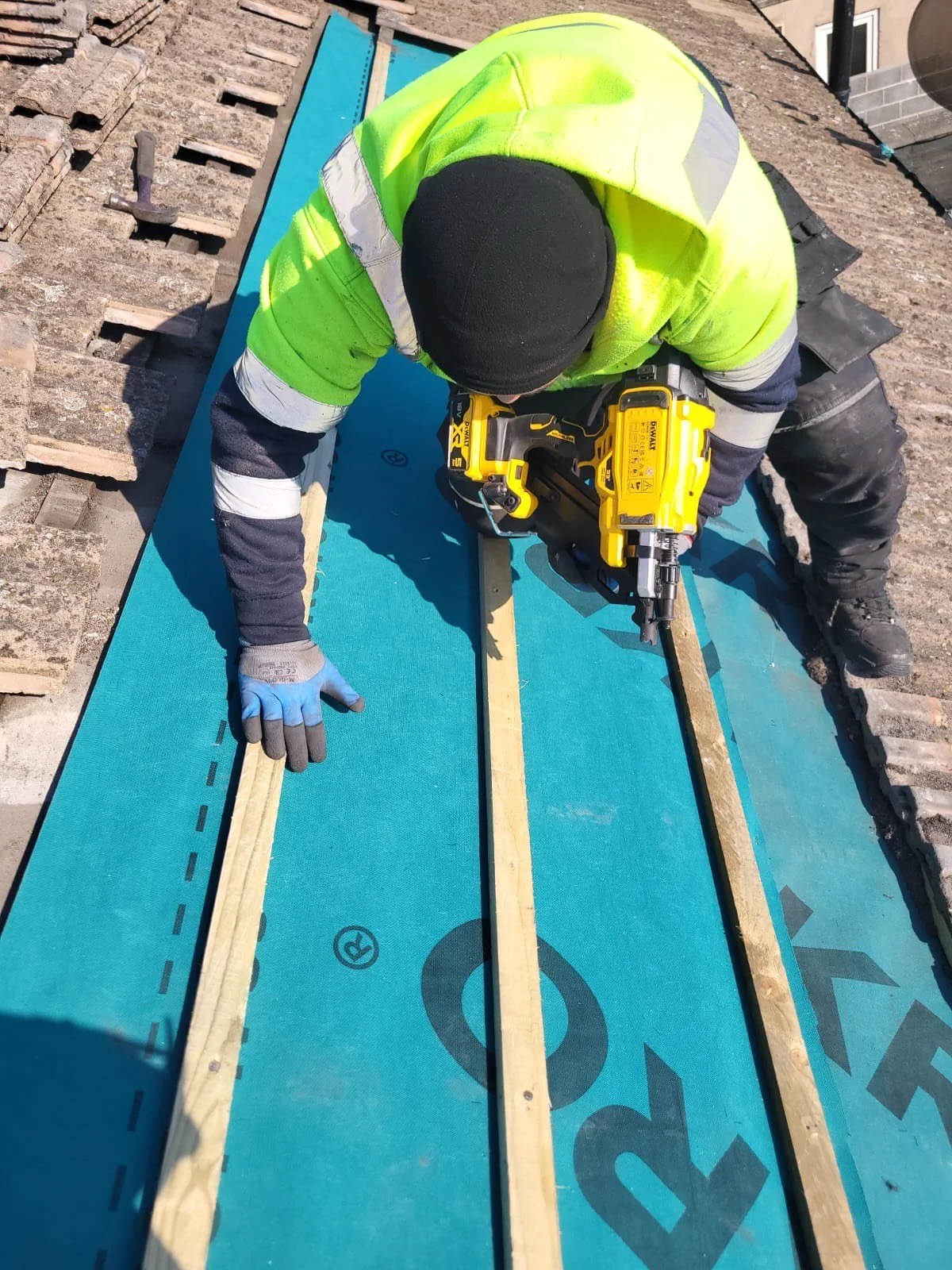 A construction worker kneeling on a blue insulation board, using a nail gun to attach wooden battens, with tools and roofing materials nearby.