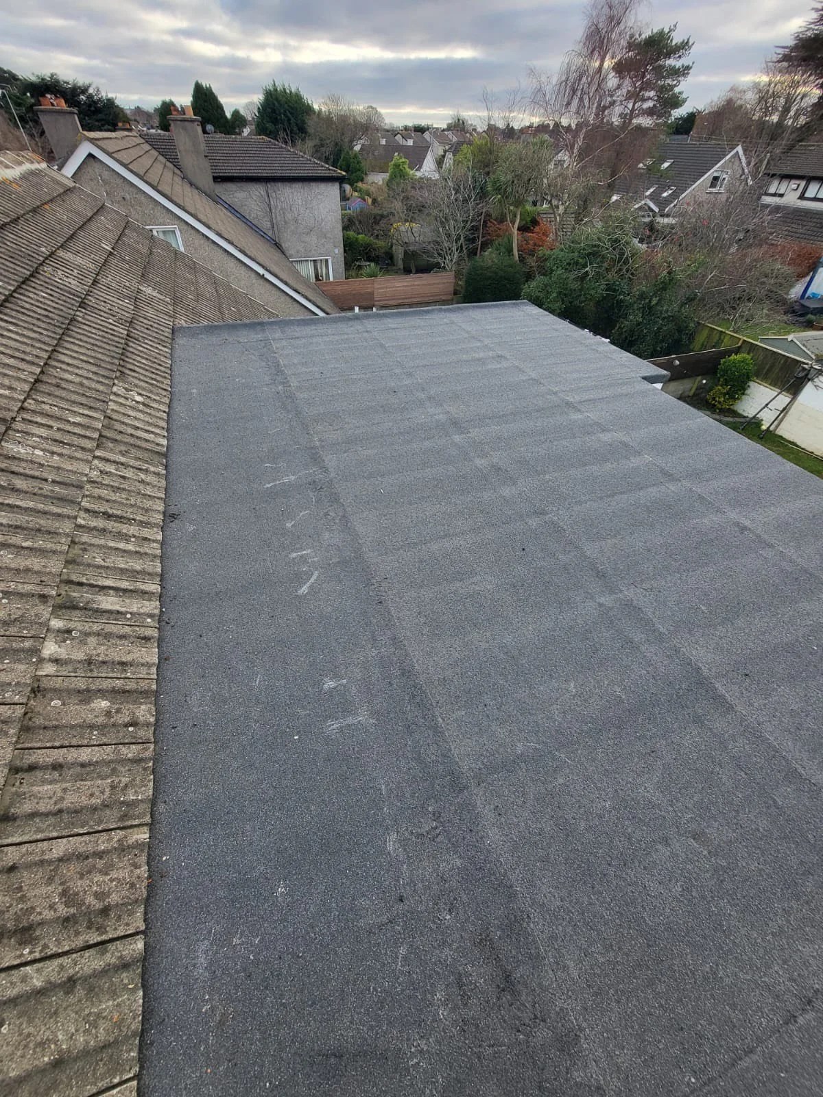 View of rooftops, trees, and neighborhood houses under a cloudy sky. In the foreground, there is a flat roofing surface next to sloped tiled roofs.