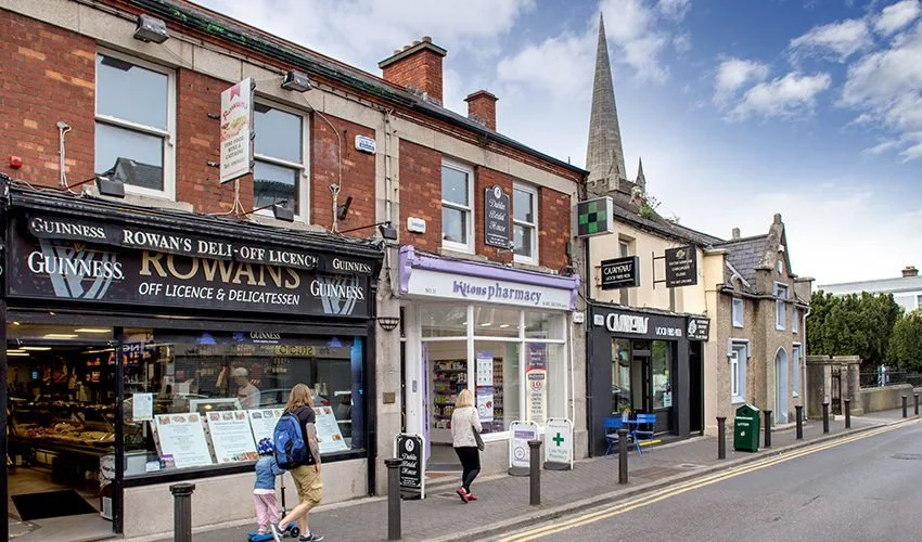 Street view of small businesses, including a deli, pharmacy, and clothing store, with pedestrians walking on the sidewalk and a church steeple in the background.