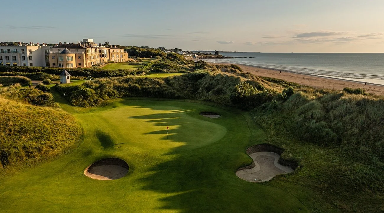 Golf course with sand bunkers near the ocean, residential buildings in the background, and a sandy beach along the shoreline at sunset.