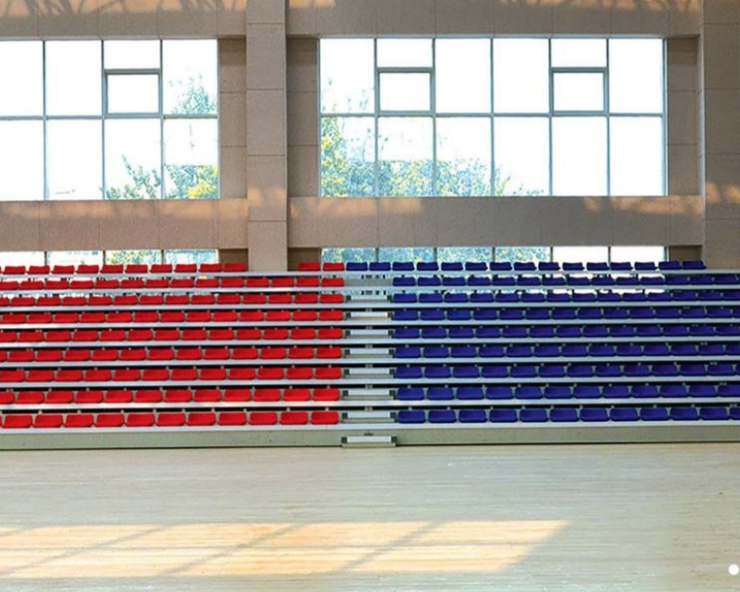 Empty indoor sports arena with red, white, and blue tiered seating, large windows in the background, and a light-colored wooden floor.