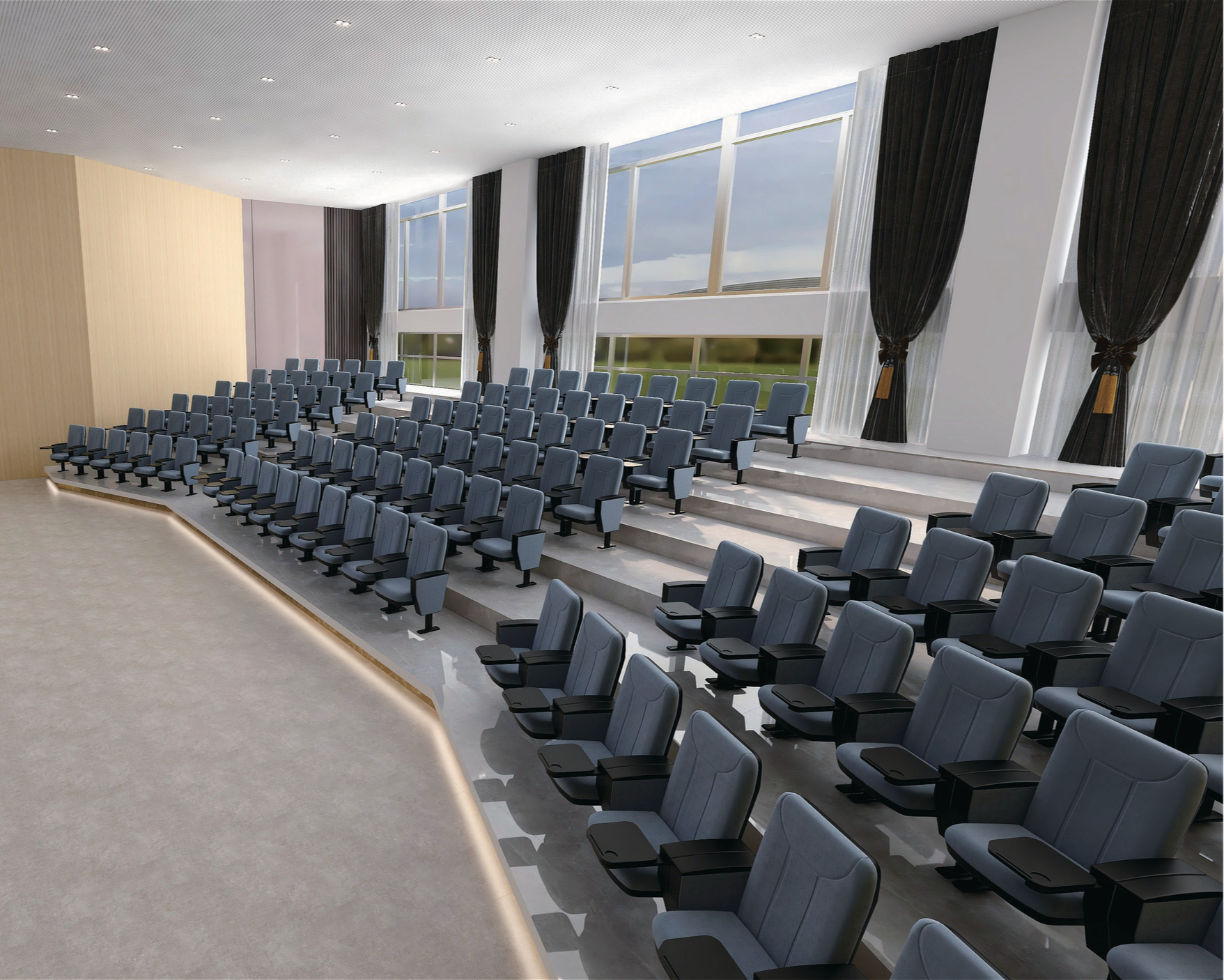 An empty large auditorium or conference room with rows of gray cushioned chairs with black armrests and attached writing surfaces, arranged in tiered seating. The room has large windows with black and white curtains, allowing natural light and a view of green landscape and cloudy sky outside. The ceiling is white with recessed lighting, and the floor is light-colored with a slight incline towards the front.