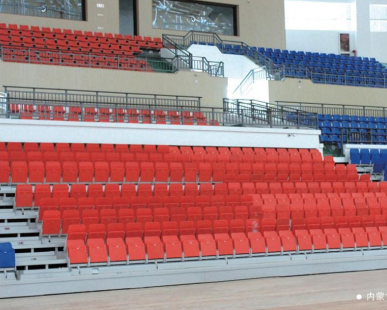 Inside an empty gymnasium with rows of red and blue bleacher seats.
