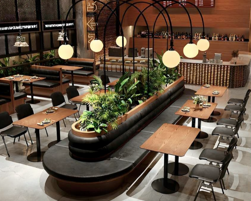 Modern restaurant interior with wooden tables, black chairs, and a curved black cushioned banquette with green plants. Hanging spherical pendant lights and a bar area in the background.
