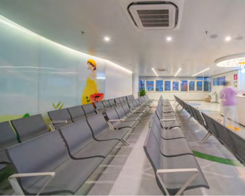 Empty waiting room with multiple rows of gray chairs, colorful wall art featuring a girl in a yellow dress holding a watermelon, and a person in a pink shirt in the background, near a reception counter.