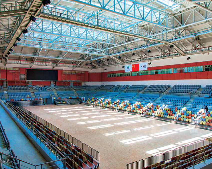 Empty indoor sports arena with colorful seats, wooden floor, and flags hanging from the ceiling.
