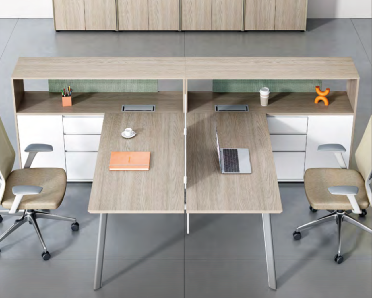A modern office shared workspace with two wooden desks pushed together, two beige swivel chairs with armrests, a shelf with colored pens, coffee cup, laptop, and small orange decorative object.