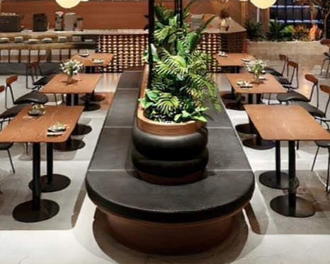 A restaurant with wooden tables and black chairs arranged around a central planter filled with green plants.