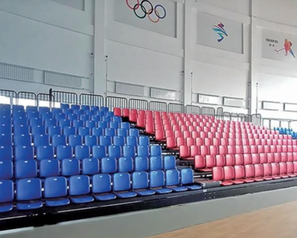 Empty blue and pink spectator seats in an indoor sports arena with Olympic and sport logos on the wall.