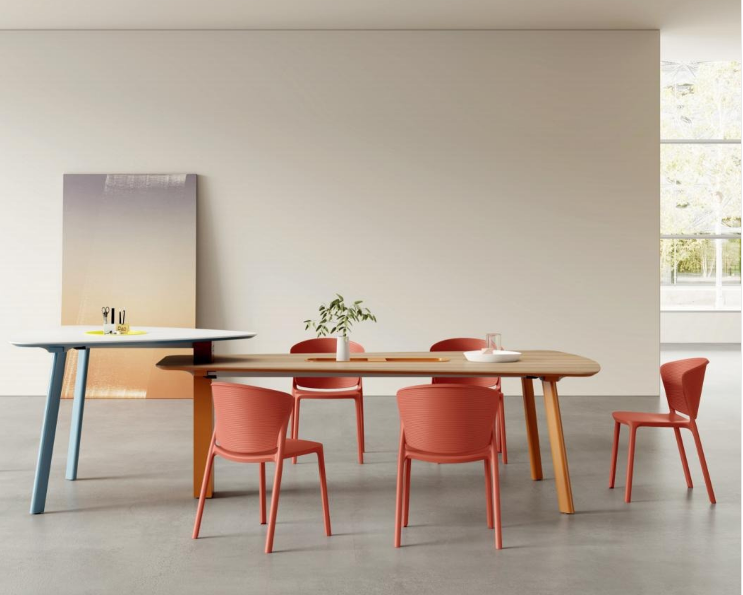 Modern dining area with a large wooden table surrounded by red chairs, a white table on the left with small office supplies, a white wall with a large abstract painting, and a window letting in natural light.