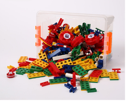 A clear plastic storage bin filled with colorful LEGO-like building bricks and pieces, with some scattered outside the bin on a white surface.