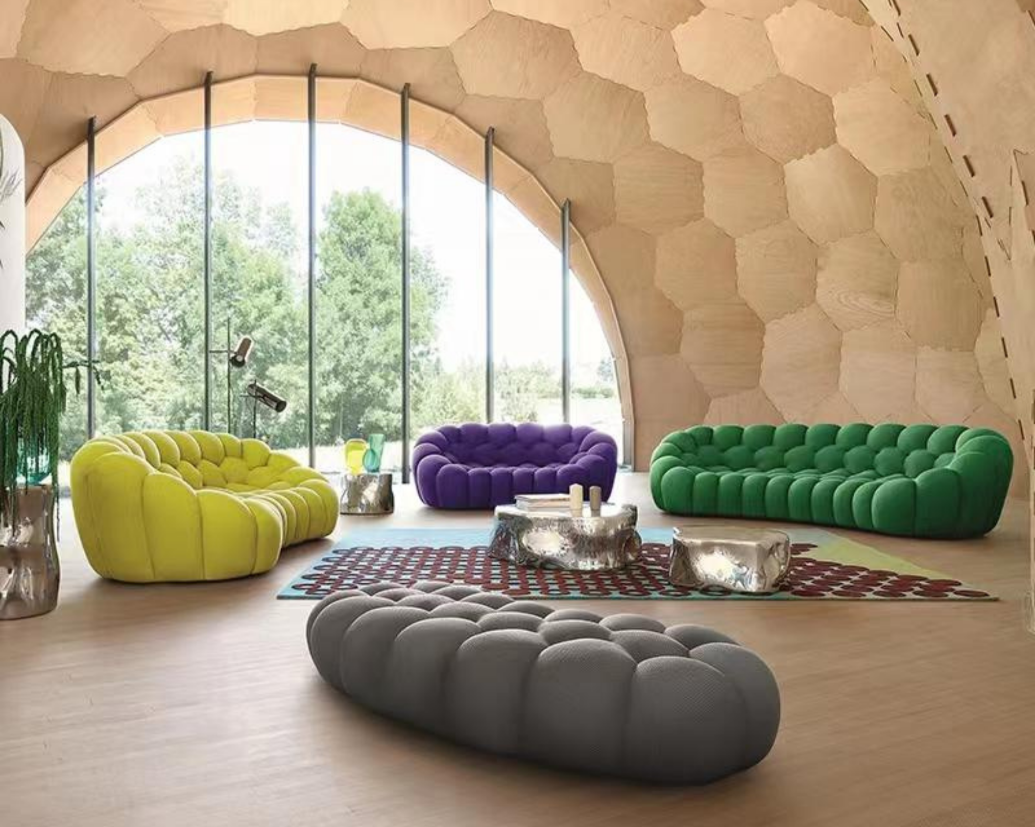 Colorful modern living room with yellow, purple, green, and gray curved velvet sofas around glass coffee tables, large floor-to-ceiling arched window showing green trees, honeycomb wooden ceiling, and wooden flooring.