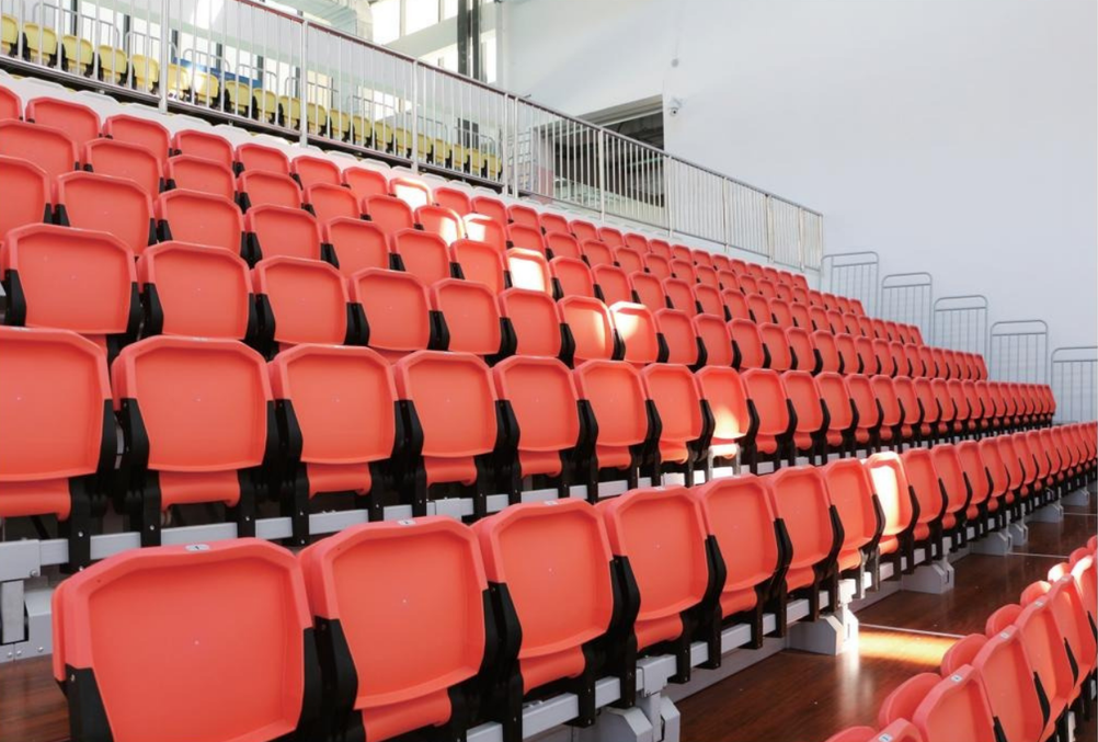 Empty orange stadium seats arranged in multiple rows inside an indoor sports arena with yellow seats visible in the background.