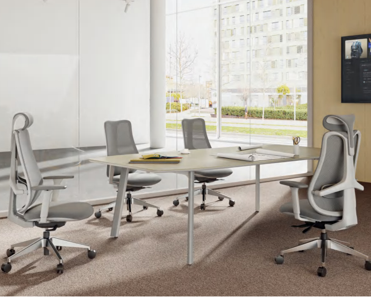 Empty modern office conference room with four gray chairs, a white table with notebooks, rolled blueprints, and a pair of scissors, large glass windows showing an outside view of a cityscape with trees and buildings.