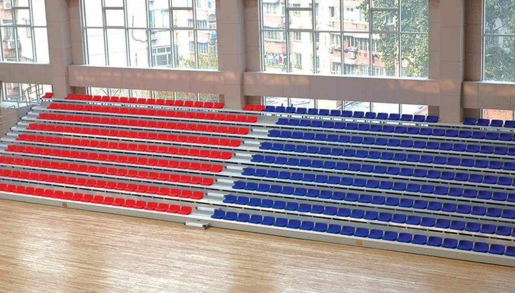 Indoor sports arena with empty red and blue bleacher seats facing large windows.