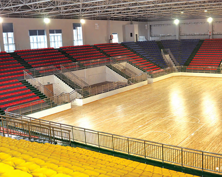 Interior view of an empty basketball gymnasium with a wooden court, colored tiered seating, and large windows.
